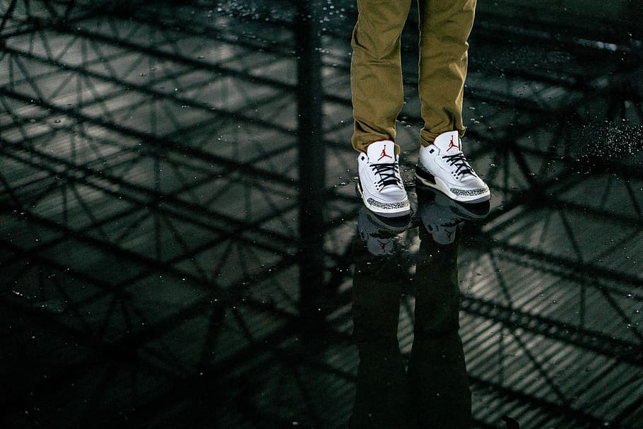 person wearing white-and-gray Air Jordan 3 standing on black tinted glass, man wearing white cement Air Jordan 3 shoes