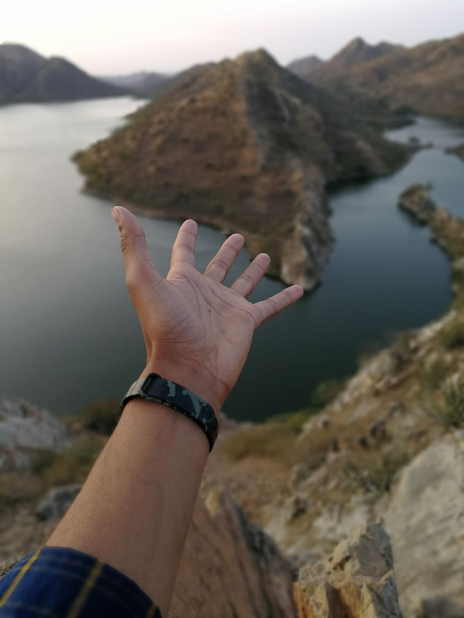 person's hand pointing to body of water and hill, person's opened hands above mountain near river HD wallpaper