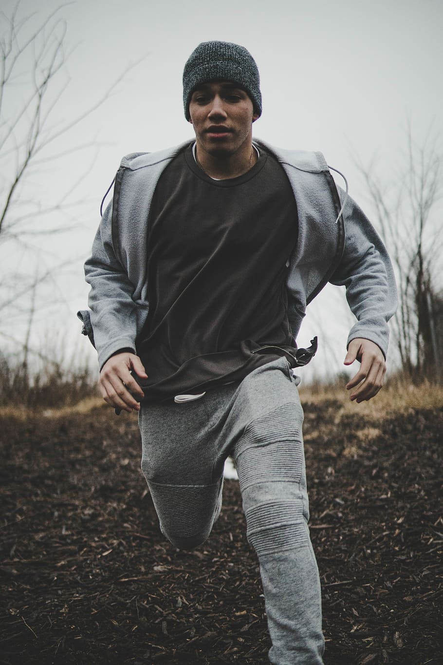 man wearing black shirt and gray jacket with pants while running into plain field, man running on brown soil near bare trees