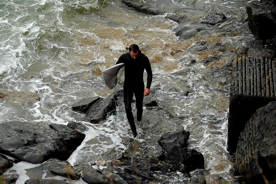 man holding white surfboard while walking on rocks, man wearing wet suit carrying surfboard HD wallpaper