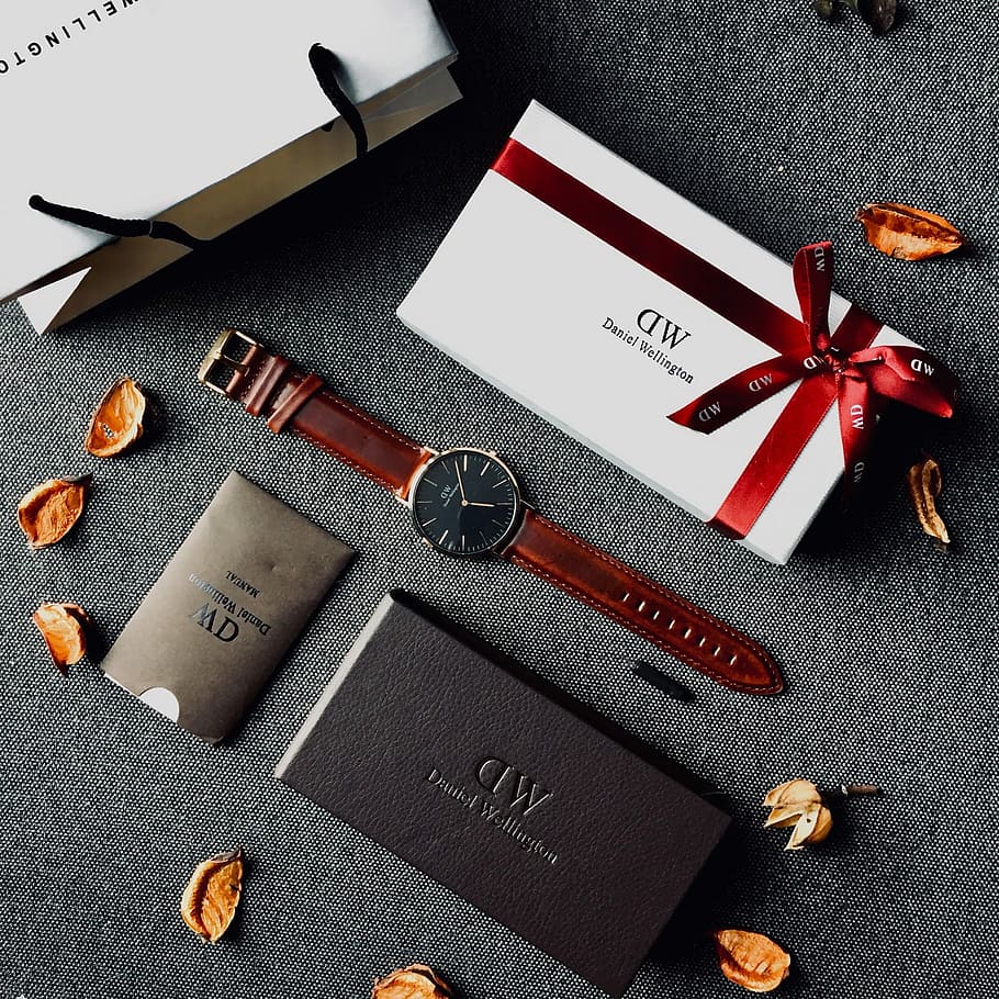 round black analog watch with brown leather stral with box and paper tote bag on gray yextile, round black analog watch with brown leather strap beside white box