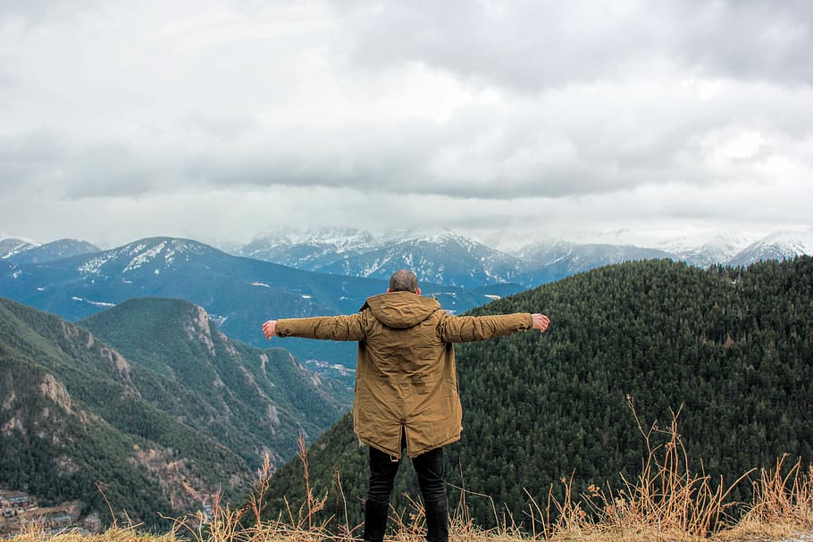 man wearing jacket standing on brown grass at the hill facing glacier mountains under white clouds during daytime HD wallpaper