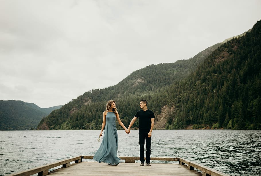 couple holding hands near body of water, man wearing black v-neck shirt beside woman wearing green maxi dress standing on brown wooden boat dock during day time