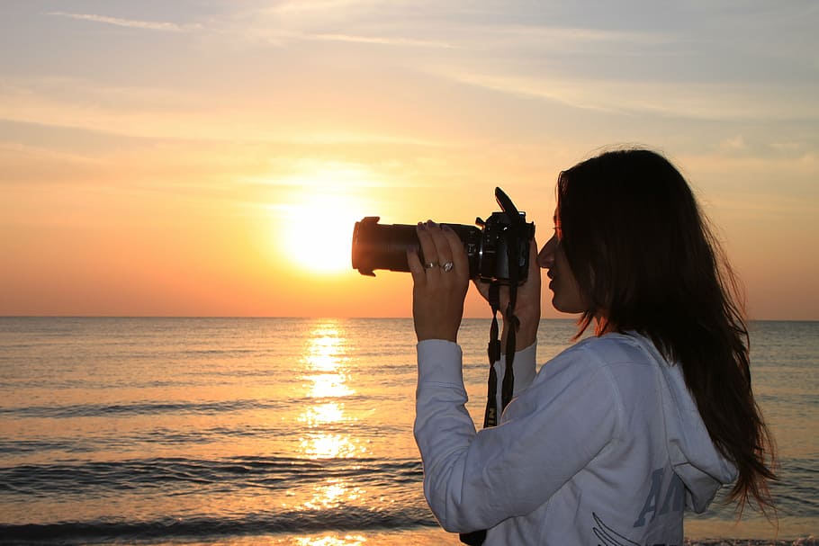 woman holding DSLR camera near beach during golden hour, female