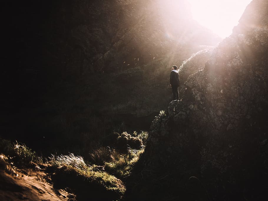 man standing on rock mountain during daytime, man standing on mountain against light at daytime HD wallpaper