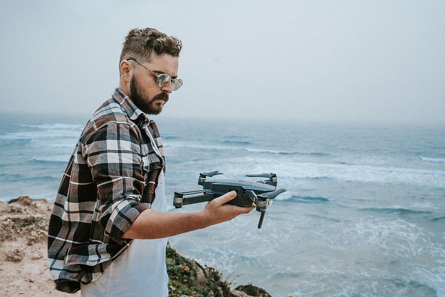 man holding black and brown DJI Mavic Pro drone on his right hand while standing on cliffside with view of sea during daytime photo, man holding black drone HD wallpaper