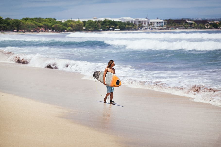 man carrying yellow and white surfboard standing near beach, surfer looking at the waves beside shore