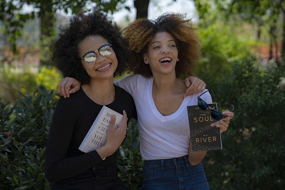 two smiling women while holding novel books, two woman side-by-side holding books in front of plants