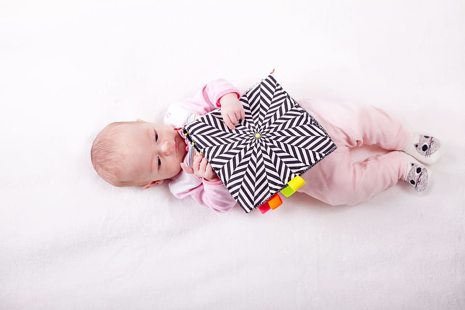 baby wearing pink pajama sleeper lying on white comforter and holding black cushion, HD wallpaper