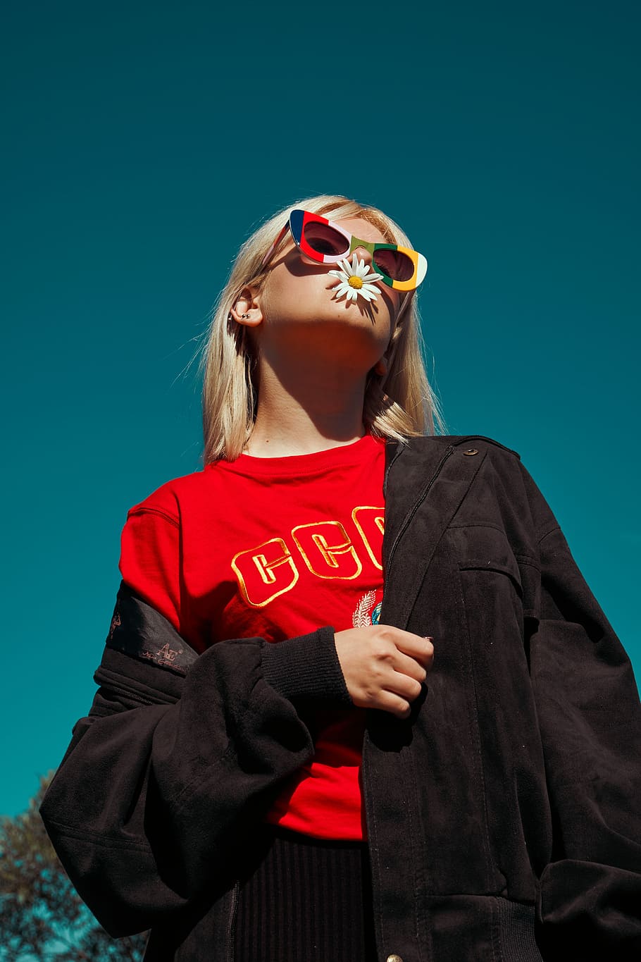 woman with sunglasses standing outdoor during daytime, low-angle photo of woman wearing red shirt and black jacket with daisy flower on mouth HD wallpaper