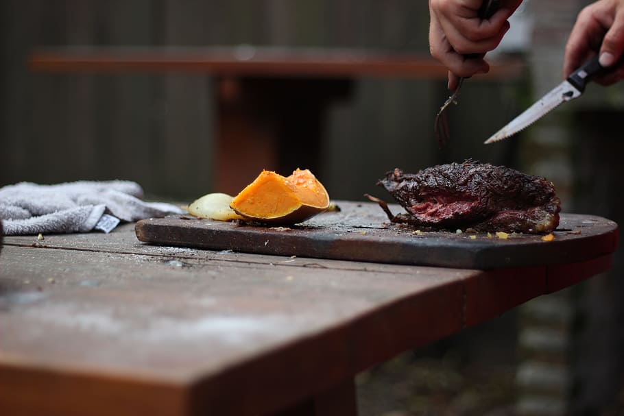 person slicing a meat using knife and fork, person slicing grilled meat near orange fruit on chopping board HD wallpaper