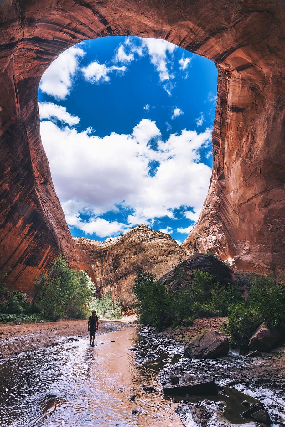 man walking under cave during daytime, man walking on body of water near arch mountain HD wallpaper