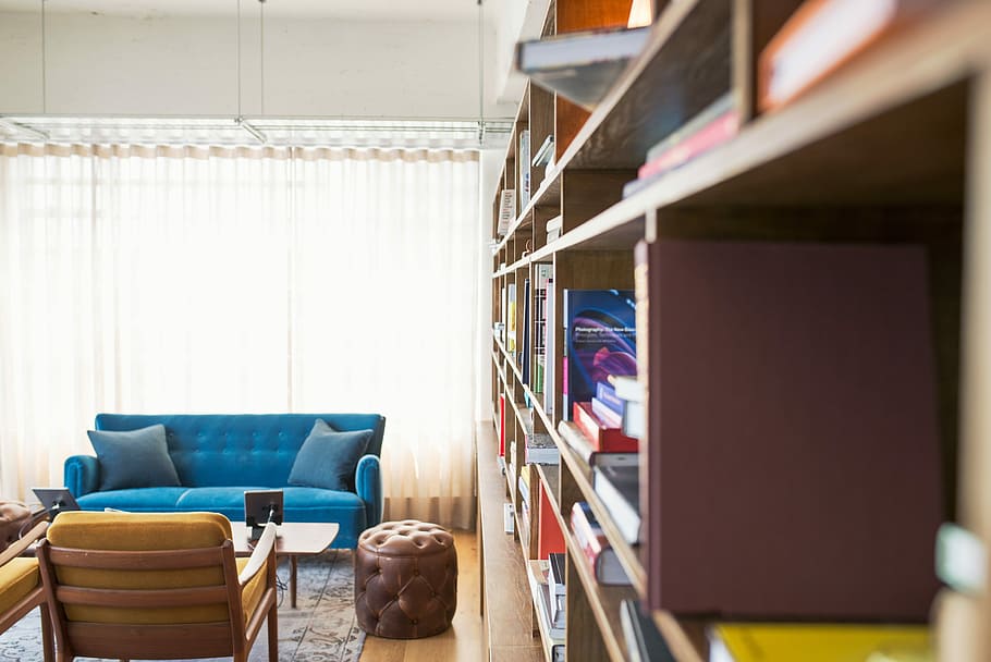 book filled wooden cabinet, tufted blue suede couch beside brown leather ottoman and brown wooden bookshelf inside room
