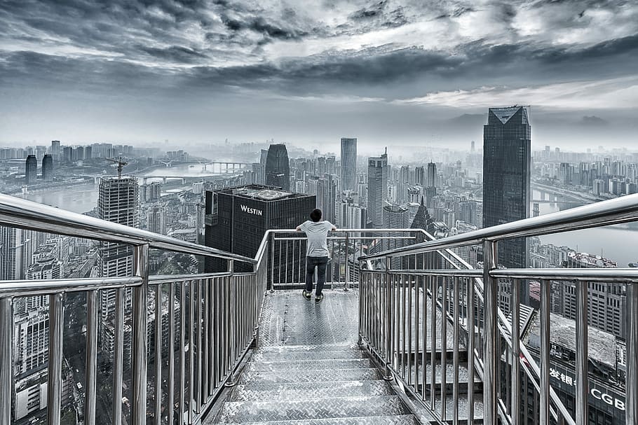man standing while hands resting on guard rail overlooking the city, man in white shirt leaning on gray stainless steel balustrade