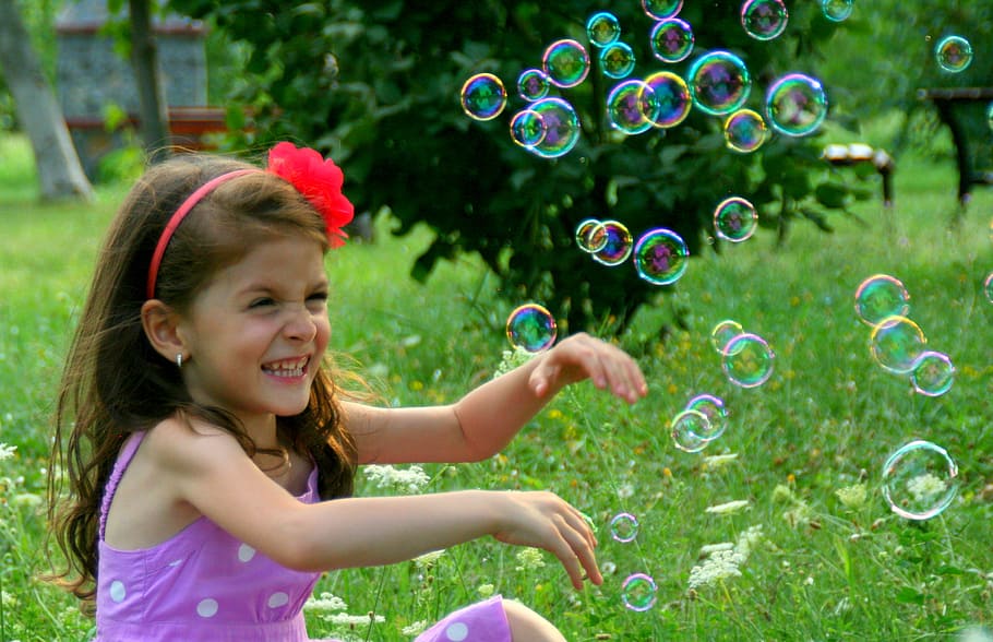 girl wearing pink dress sitting on grass near bubbles, soap bubbles