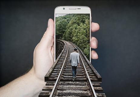 man walking on train rail through person holding white smartphone optical illusion artwork HD wallpaper
