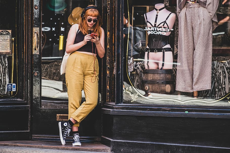 woman standing in front of store while using a smartphone, woman standing in front of dress shop while wearing headphones HD wallpaper