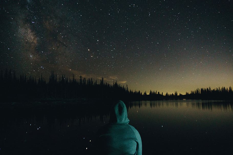person wearing green hoodie facing on body of water and trees, person sitting in front of body of water under night sky