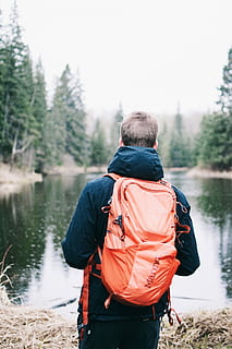 man wearing blue jacket and red backpack facing body of water, man with bag in front of lake near trees HD wallpaper