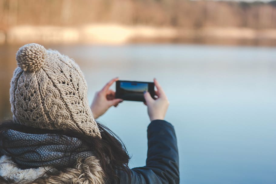 woman in black leather jacket holding smartphone taking picture of body of water HD wallpaper