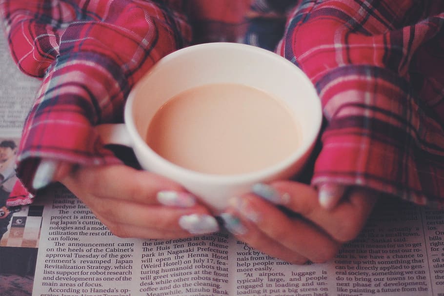 person in red tops touching filled white ceramic mug on top of newspaper