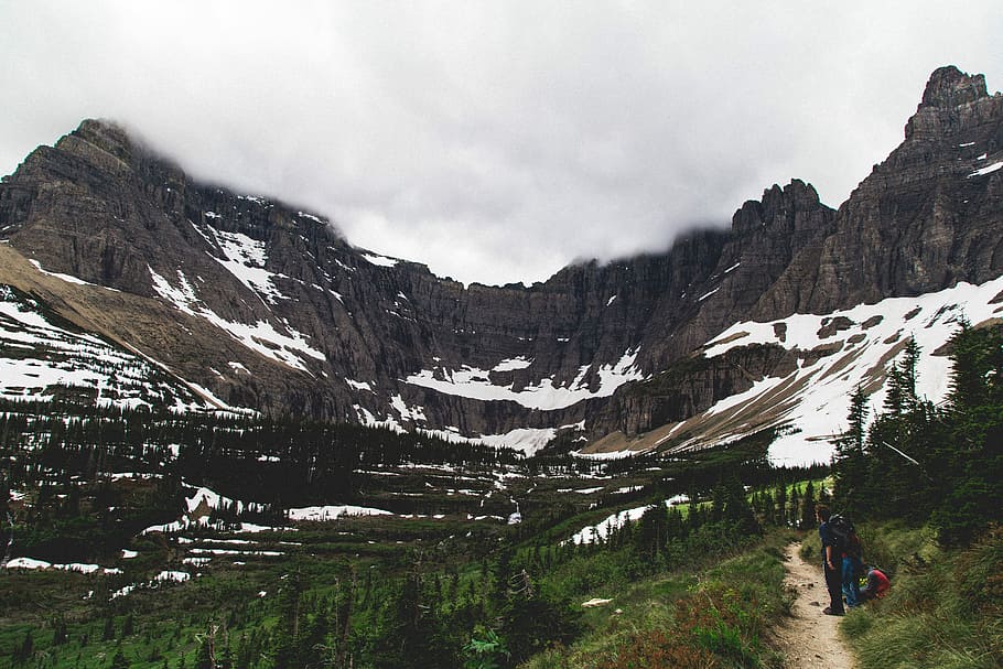 three people hiking on hill with snow caped mountain in distance, person standing near mountain during daytime HD wallpaper
