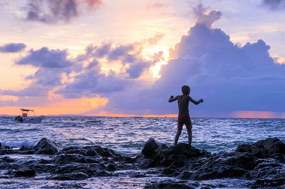 silhouette of man standing on rock formations near body of water HD wallpaper