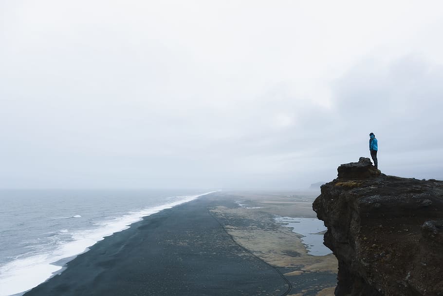 person on hilltop during daytime, person standing on cliff in front of ocean during daytime