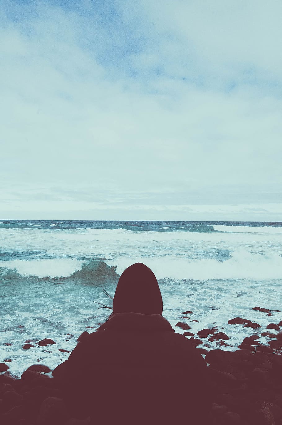person sitting on rocking shore under cloudy sky, person facing boulders and body of water HD wallpaper