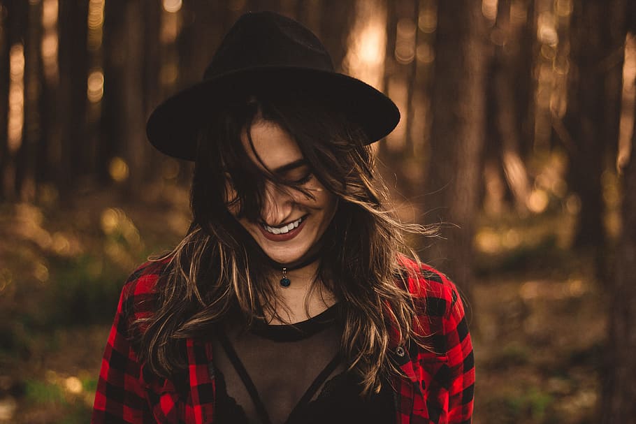 woman in fedora hat surrounded by trees, woman smiling under shade of trees in woodlands at daytime HD wallpaper