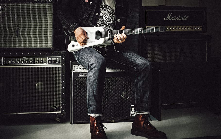 man wearing blue denim jeans playing white and black electric guitar sitting on black guitar amplifier, man holding electric guitar near speakers