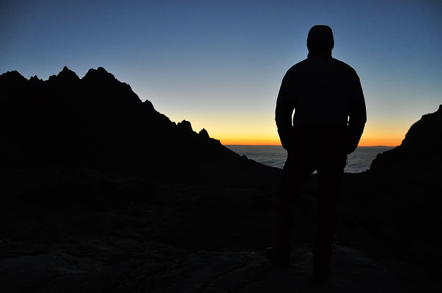silhouette of person on top of mountain under blue sky during orange sunset, silhouette man standing near sea during sunset