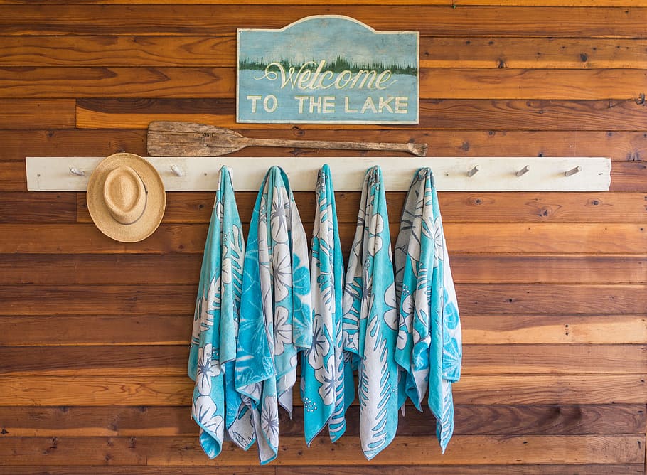 brown hat in wall hook, white-and-teal floral towels on white wooden rack with Welcome to the Lake signage HD wallpaper