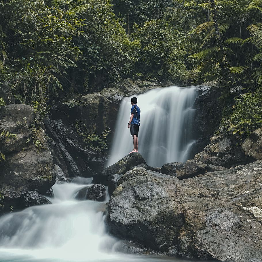 man standing on rocks with waterfalls background, man in blue top standing on rock beside of water falls during daytime, HD wallpaper