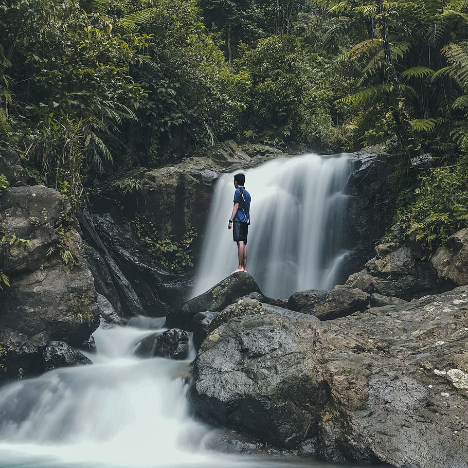 man standing on rocks with waterfalls background, man in blue top standing on rock beside of water falls during daytime HD wallpaper