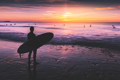A surfer stands on the beach and looks out to the ocean at sunset. Image captured in Cornwall, England HD wallpaper