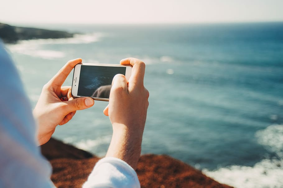 person standing on cliff taking photo of body of water during daytime, person holding opened Android smartphone taking photo of body of water HD wallpaper