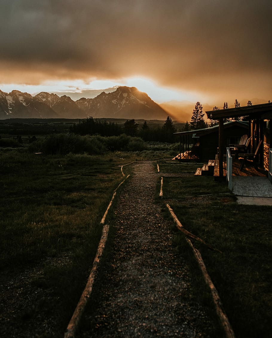 wooden house near mountain during golden hour, overlooking view of mountains under orange sky and dark clouds HD wallpaper