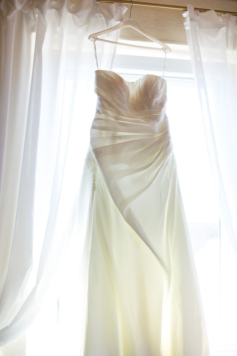 white drape tube wedding gown hang on clothes hanger against window curtains