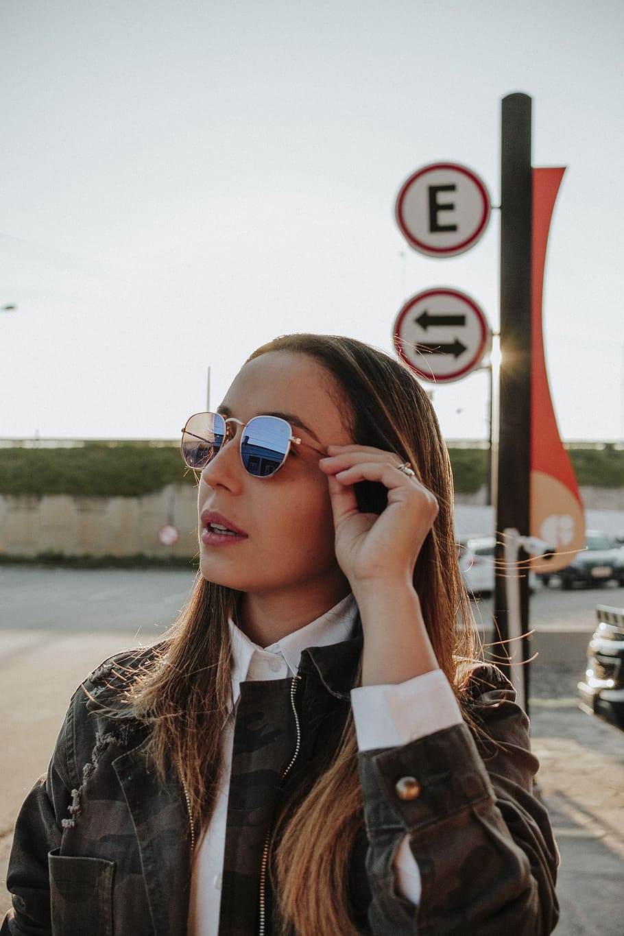 woman holding her sunglasses standing on parking lot during daytime, woman wearing green, beige, and black camouflage jacket with eyeglasses near signage, HD wallpaper