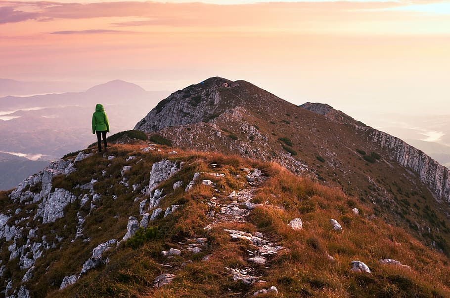 person wearing green jacket standing on cliff facing mountain, person wearing green bubble jacket standing over rocky cliff under cloudy sky HD wallpaper
