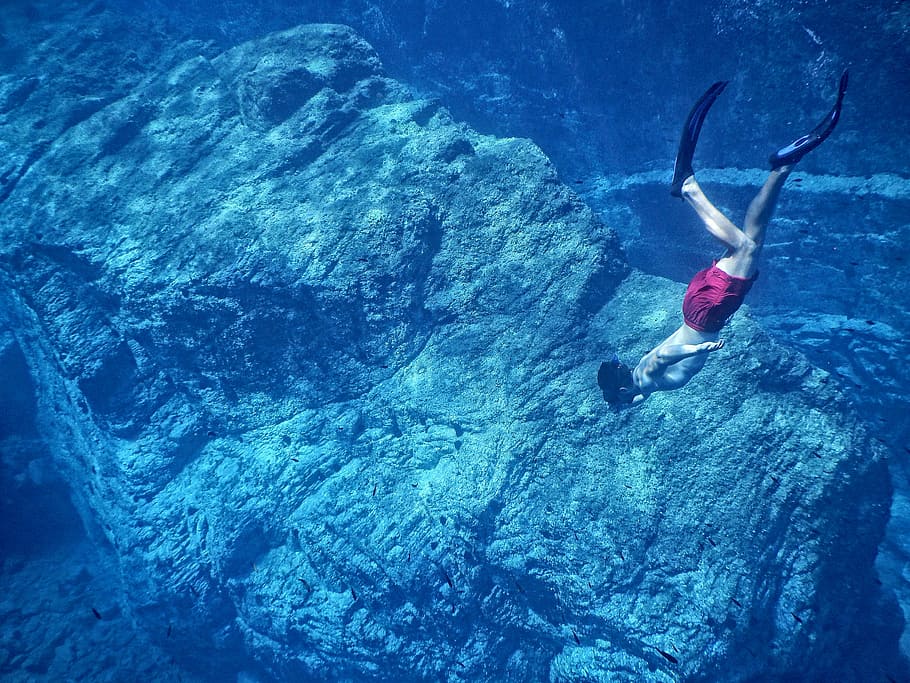 man in red shorts swimming near huge underwater rock, man free diving on body of water