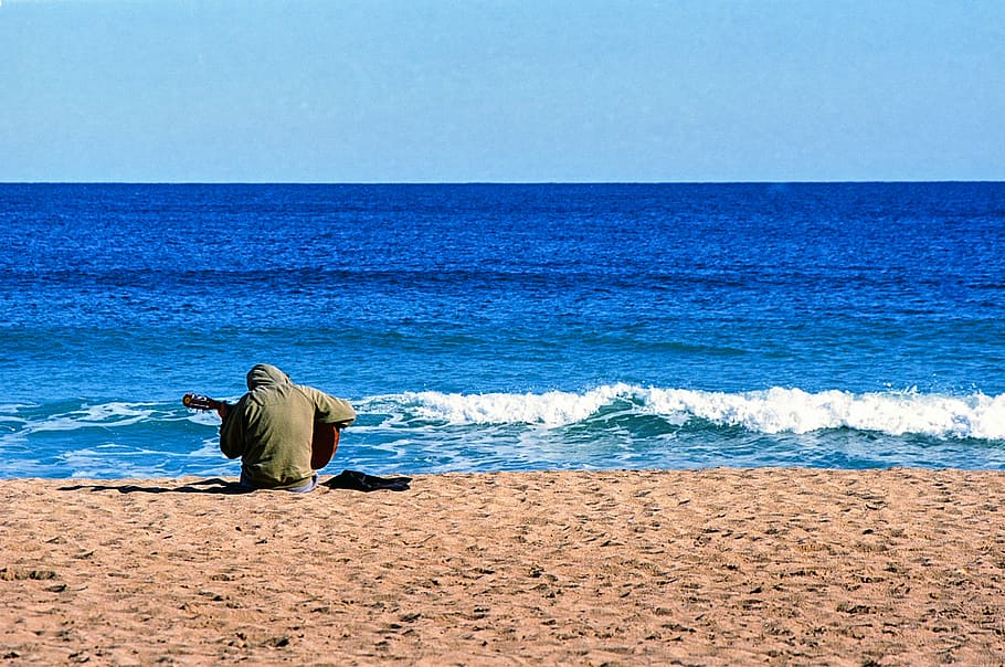 man holding guitar sitting on sand near body of water, Beach