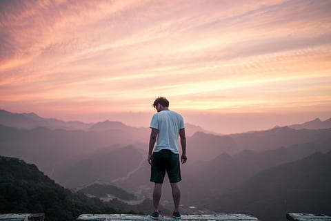 man standing on cliff, man standing in front of mountain during daytime HD wallpaper