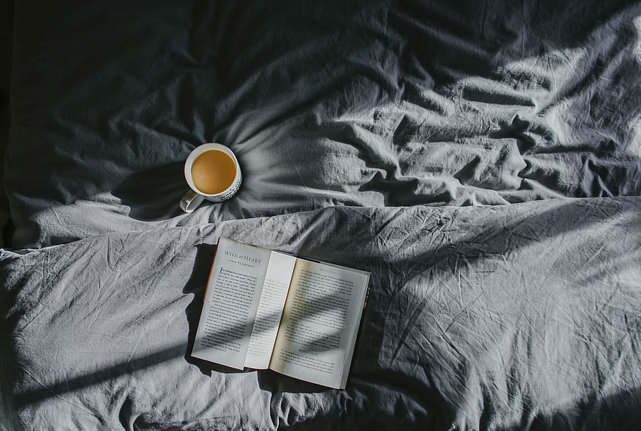 white ceramic mug beside book on gray textile, open book near mug on bed cover HD wallpaper