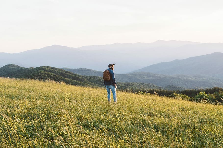 man standing on grass field in mountain, man standing on green grass field facing mountain during daytime HD wallpaper