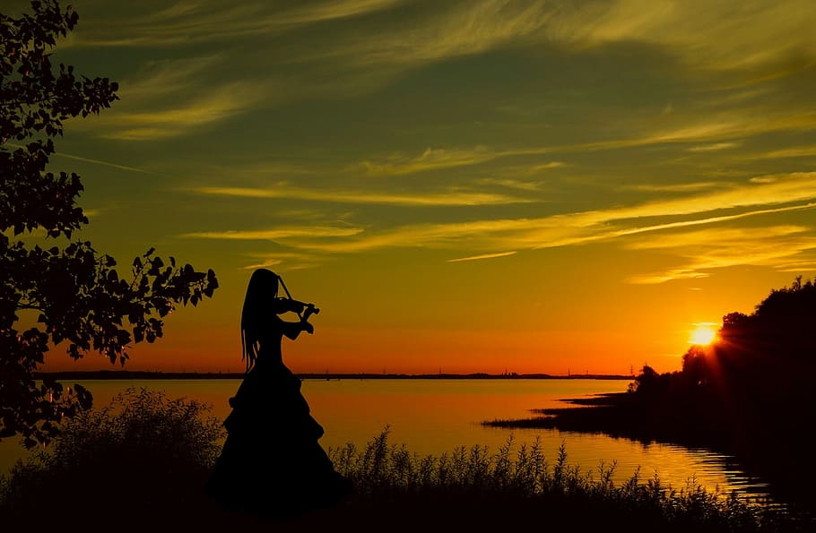 silhouette of woman playing violin beside water during golden hour