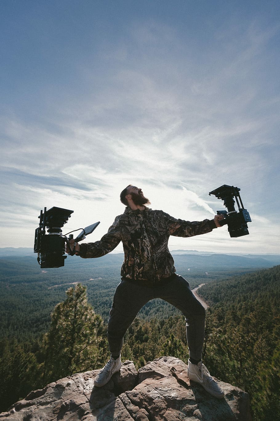 person standing on rocky mountain peak while holding two cameras, person standing on rock while holding device near trees at daytime HD wallpaper