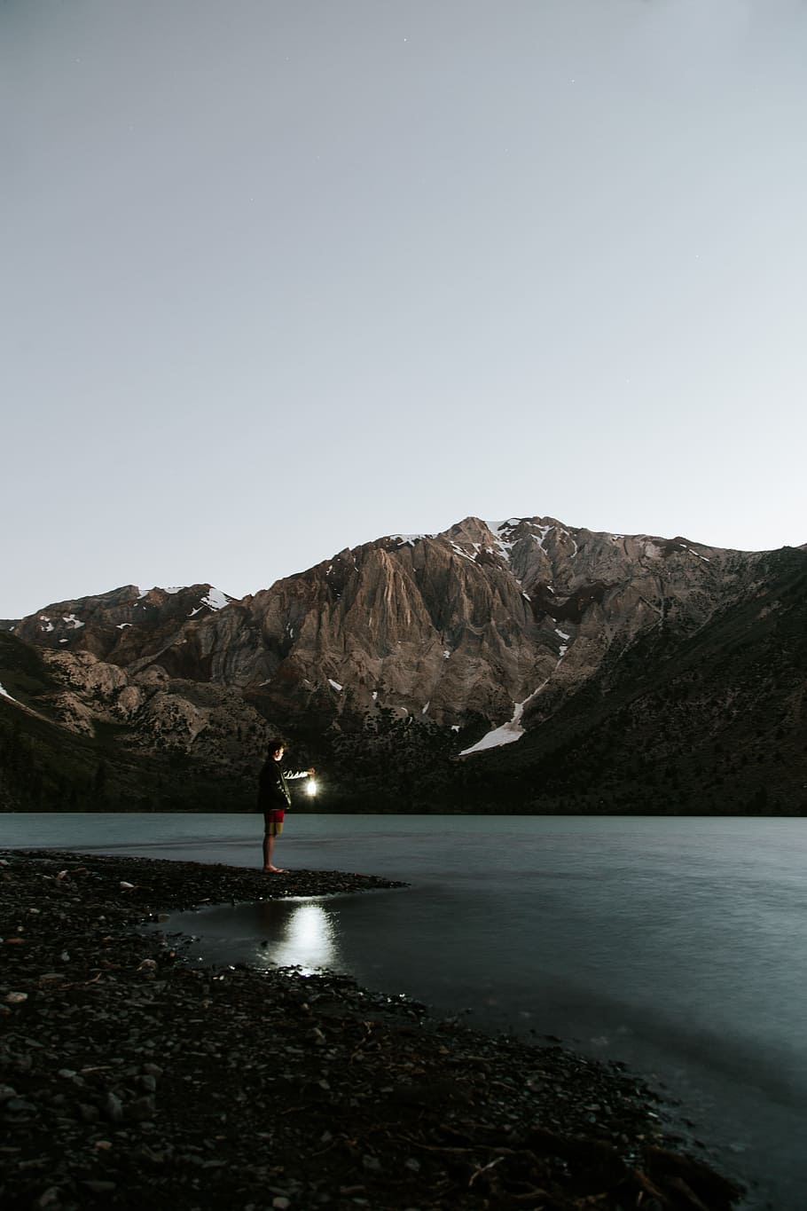 man near body of water, man standing near shore across the brown mountain with cloudy sky during daytime HD wallpaper