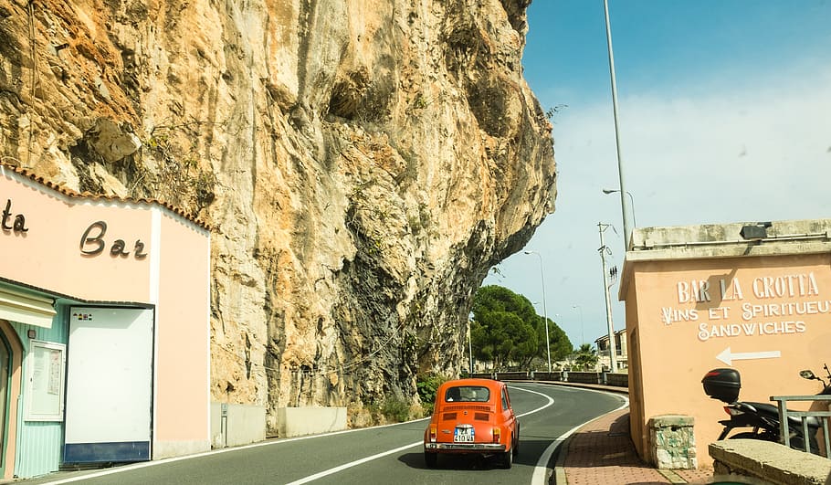 red Volkswagen Beetle coupe on black concrete road between concrete buildings under white and blue sky at daytime, red vehicle on gray concrete road between brown concrete building and brown rock formation during daytime HD wallpaper